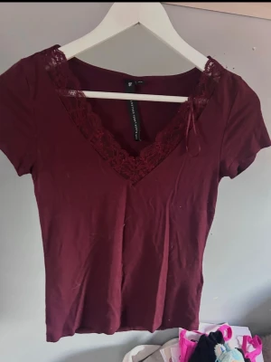 Vinröd topp med spetsdetalj - Snygg vinröd topp med v-ringning och spetskant runt halsen. Kortärmad och figurnära passform, perfekt att styla med jeans eller kjol. Tillverkad i mjukt material som känns skönt mot huden.