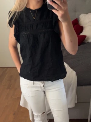 Svart blus med volangdetaljer - Säljer en svart blus med volangdetaljer på axlarna och diskreta spetsinslag. Blusen har en lös passform och är kortärmad, perfekt för en trendig och avslappnad look.