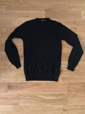Svart stickad tröja från J.Lindeberg - Klassisk svart stickad tröja från J.Lindeberg med rund halsringning och ribbade muddar vid ärmslut och nederkant. Enkel och stilren design som passar till det mesta. Perfekt basplagg för garderoben. Priset ej hugget i sten!