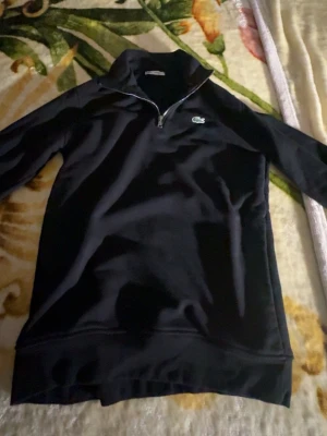 Svart zip hoodie från Lacoste - Snygg svart hoodie från Lacoste med dragkedja halvvägs ner och klassiska krokodilloggan på bröstet. Tillverkad i mjukt material och har hög krage. Perfekt för dig som vill ha en stilren och bekväm look. Använt 2 gånger för stor 