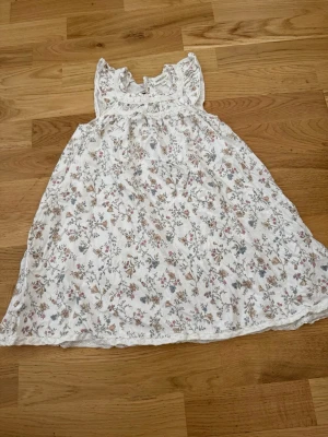 Vit blommig klänning från Newbie - Söt vit klänning från Newbie med blommigt mönster i pastellfärger. Klänningen har spetsdetaljer vid ärmar och nederkant samt ärmlös design. Perfekt för sommarens varma dagar.