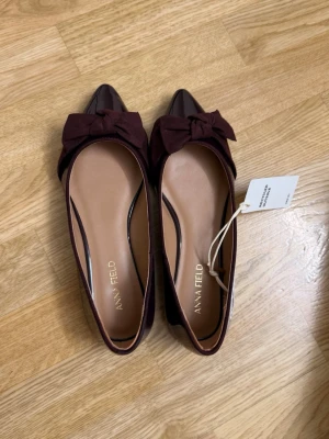 Vinröda ballerinaskor med rosett - Snygga ballerinaskor från Anna Field i vinrött lack och mockaimitation. Skorna har spetsig tå och en stor rosett framtill som ger en trendig touch. Perfekta för dig som vill ha något extra till outfiten. Fodrade med beige innersula för skön känsla.