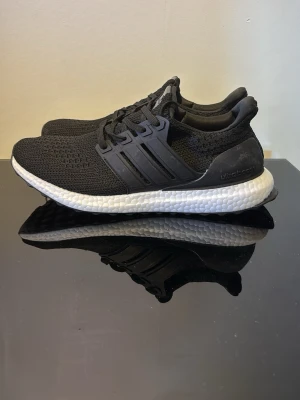 Svarta Adidas Ultraboost sneakers - Säljer ett par svarta Adidas Ultraboost sneakers med stickad ovandel och vita Boost-mellansulor. Skorna har de klassiska tre ränderna på sidan och svart yttersula med Continental-gummi för extra grepp. Perfekta för dig som gillar stilrena och bekväma sneakers.