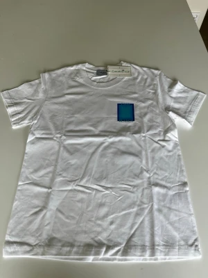 Vit t-shirt med blått tryck Casablanca - Säljer en vit t-shirt från Casablanca med ett blått grafiskt tryck på bröstet och en stor blå rektangel med texten 'CASA BLANCA' på ryggen. Klassisk passform och rund hals. Perfekt för dig som gillar stilrena men ändå unika plagg.
