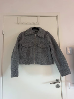 Grå teddyjacka med dragkedja - Säljer en grå teddyjacka från Zara med dragkedja framtill och två stora bröstfickor med lock. Jackan har en bred krage och är tillverkad i ett mjukt, fluffigt material som ger en mysig look. Perfekt för kyliga dagar och enkel att styla till olika outfits. Använd fåtal gånger