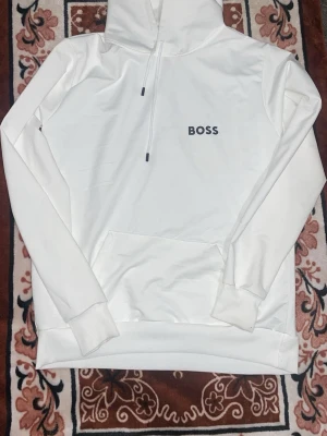 Vit hoodie från BOSS med tryck - Snygg vit hoodie från BOSS med svart logotyp på bröstet. Hoodien har dragsko i huvan och en stor magficka framtill. Tillverkad i mjukt material som känns skönt mot huden. Perfekt för en clean och stilren look.