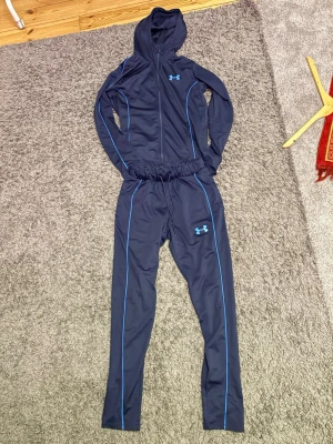 Marinblå träningsset från Under Armour - Snyggt marinblått träningsset från Under Armour med matchande hoodie och byxor. Setet har ljusblå detaljer och logga på både jacka och byxa. Tillverkat i ett stretchigt funktionsmaterial som passar perfekt till träning eller uppvärmning.