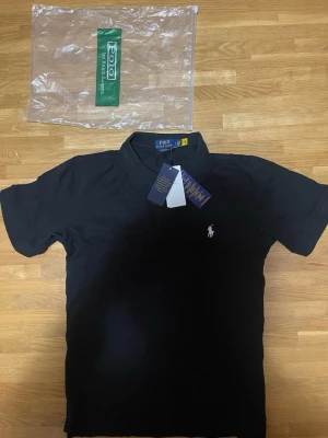 Svart pikétröja från Polo Ralph Lauren - Klassisk svart pikétröja från Polo Ralph Lauren med vit broderad logga på bröstet. Tröjan har krage, korta ärmar och knappar vid halsen. Perfekt för en stilren och enkel look.