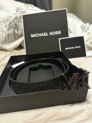 Svart Michael Kors bälte med MK-spänne - Snyggt svart bälte från Michael Kors med stort MK-spänne i metall. Bältet har diskret mönster längs hela bandet och levereras i originalask. Perfekt accessoar för att lyfta din outfit.