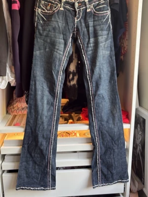 Mörkblå bootcut jeans med kontrastsömmar - Säljer ett par mörkblå bootcut jeans från Laguna Beach med vita och rosa kontrastsömmar. Jeansen har dekorativa knappar med liljemotiv, slitna detaljer och flera fickor fram. Perfekta för dig som gillar en unik och edgy stil. Unika fickor bak som liknar både Miss Me men även True Religion. Min mammas gamla vintage jeans. Storlek 26 men är lite tajta.