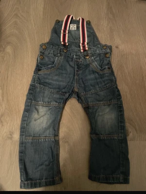 Jeansjumpsuit med hängslen från Boxing - Jeansjumpsuit i blå denim med vita, röda och marinblå randiga hängslen. Byxan har flera dekorativa sömmar och fickor framtill samt bruna knappar. Hängslena är justerbara med metallspännen och ger en cool retrostil. 18-24 månader 