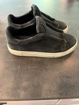 Svarta sneakers från Axel Arigato - Snygga svarta sneakers från Axel Arigato i mocka med vit sula. Modellen har en stilren design utan snörning och diskret logga på sidan. Perfekta för dig som gillar minimalistisk och modern stil.