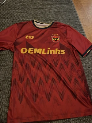 Röd fotbollströja Spain OEMLinks - Säljer en röd fotbollströja med gult tryck från Spain med OEMLinks-logga på bröstet. Tröjan har korta ärmar, svarta och gula detaljer vid hals och ärmslut samt ett mönster med svarta zigzag-linjer framtill.