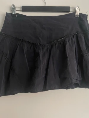 Svart volangkjol i bomull - Snygg svart kjol med volangdetaljer och lätt rynkade partier. Kjolen är kort och har en avslappnad passform, perfekt för en trendig och ungdomlig stil. Tillverkad i ett mjukt bomullstyg som känns bekvämt mot huden.