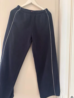Low waist mörkblå mjukisbyxor med vita ränder - Low waist mörkblå mjukisbyxor med vita kontrastsömmar längs benen. Byxorna har resår i midjan och raka ben för en avslappnad look. Perfekta för chill dagar eller när du vill vara bekväm. Materialet känns mjukt och skönt mot huden.