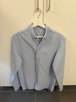 Ljusblå skjorta från H&M - Klassisk ljusblå skjorta från H&M i regular fit. Skjortan har knappar framtill, bröstficka och button down-krage. Tillverkad i mjuk bomull som känns skön mot huden. Perfekt för en clean och stilren look.