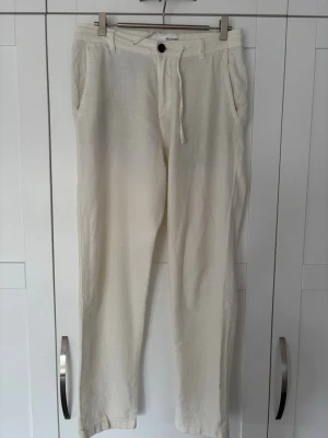 Vita linnebyxor från Selected Homme - Säljer ett par vita linnebyxor från Selected Homme med avslappnad passform, dragsko i midjan och sidofickor. Byxorna har raka ben och två bakfickor med knapp. Perfekta för en chill och stilren look under varma dagar.