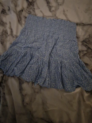 Blå blommig volangkjol - Söt blå kortkjol med små vita blommor och volangkant. Kjolen har en smockad midja som ger en bekväm och flexibel passform. Perfekt för dig som gillar en feminin och somrig stil.