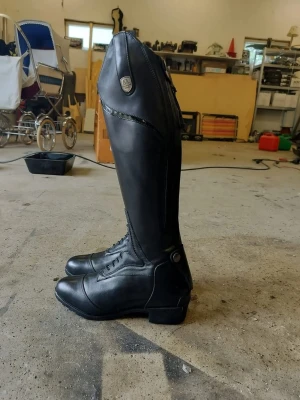 Svarta ridstövlar från Mountain Horse - Säljer ett par svarta ridstövlar från Mountain Horse i slätt läder. Stövlarna har snörning framtill, dragkedja bak och en liten knappdetalj vid ankeln. Klassisk och stilren modell med hög skaft och diskret logga på sidan. Aldrig använda. Originellt pris är 3000.