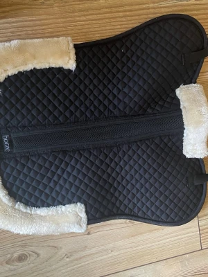 Svart fårskinnspadd från Horze - Säljer en svart padd från Horze med quiltat mönster och mjukt beige syntetfårskinn längs kanterna. Perfekt för att lägga under sadeln för extra komfort till hästen. Materialet är slitstarkt tyg och syntetpäls. Storlek m/L  cob/ full. Helt ny