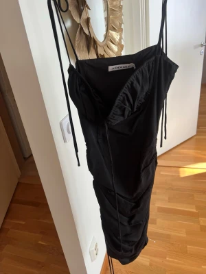 Svart klänning från Adoore - Snygg svart klänning från Adoore med tunna axelband och omlottdetalj vid bysten. Klänningen har en figurnära passform och är tillverkad i ett mjukt material som ger en elegant look. Perfekt för dig som vill ha en stilren och trendig outfit.