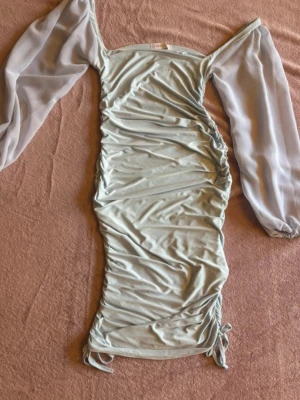 Ljusblå klänning Femme Luxe - Trendig ljusblå kortklänning från Femme Luxe och långa, transparenta ärmar. Klänningen har snygg snörning i sidorna och är tillverkad i ett mjukt, stretchigt syntetmaterial. Perfekt för dig som vill sticka ut med en unik look.