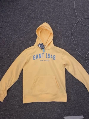 Gul Gant Hoodie. - Gul Gant Hoodie med texten Gant 1949, New Haven på tröjan. En väldigt fin Hoodie med skön och mycket bra kvalitet. Hoodien är aldrig använd och säljs pågrund av att den är för liten. Den har prislappen kvar som du ser. Ny pris 800kr. Kvalitet 78% bomull och 22% polyester. Storlek 158/164!