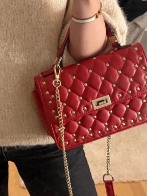 Röd quiltad handväska med nitar - Trendig röd handväska med quiltat mönster och guldfärgade nitar. Väskan har en guldkedja som axelrem och ett vridlås framtill. Perfekt accessoar för att lyfta din outfit med en edgy touch.