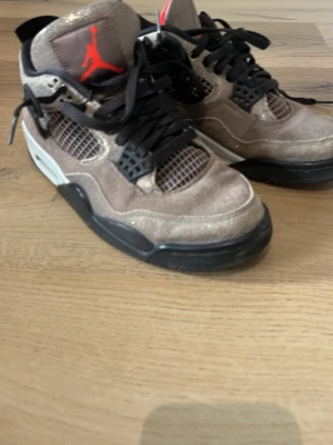 Air Jordan 4 Taupe Haze sneakers med box - Snygga Air Jordan 4 Taupe Haze sneakers med brun mocka, svarta detaljer och meshpaneler. Klassisk Jumpman-logga i rött på plösen och svarta skosnören. Tjock sula i svart och vitt ger en sportig vibe. Perfekta för dig som gillar streetwear och vill sticka ut.