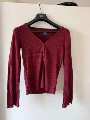 Vinröd tröja  - Vinröd ribbad cardigan från GAP med v-ringning och knappar framtill. Långärmad modell i mjukt stretchigt material som sitter snyggt på kroppen. Perfekt att styla med både jeans och kjol för en trendig look.