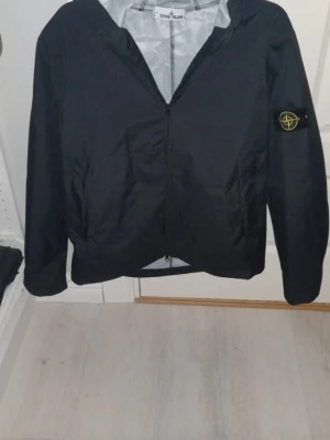 Svart vindjacka från Stone Island - Snygg svart vindjacka från Stone Island med huva och dragkedja framtill. Jackan har den klassiska logotypen på vänster ärm och två sidofickor. Tillverkad i lätt och vindtätt material, perfekt för blåsiga dagar.