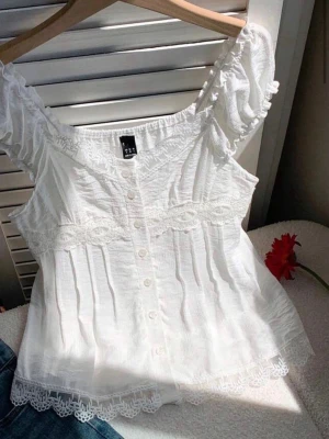 Vit spetsprydd blus - Söt vit blus från Shein med spetsdetaljer och knappar framtill. Blusen har korta puffärmar, vacker spetskant nedtill och runt midjan samt en luftig, lätt struktur. Perfekt för en somrig look med romantiska vibbar.