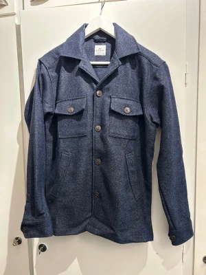 Mörkblå overshirt med 4 fickor - Stilren mörkblå overshirt i kraftigt tyg med två bröstfickor med lock och knappar samt sidofickor. Klassisk krage och stora bruna knappar framtill och vid ärmslut. Perfekt lager på lager-plagg för en avslappnad look. Sparsamt använd.