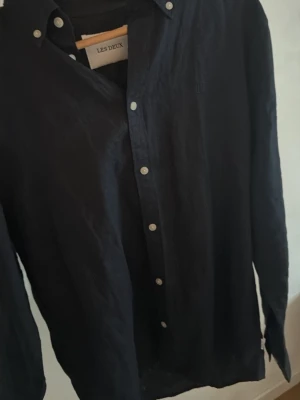 Svart linneskjorta från Les Deux - Stilren svart skjorta från Les Deux i linne med klassisk krage och vita knappar framtill. Skjortan har lång ärm och diskret broderad logga på bröstet. Perfekt för en clean och avslappnad look nypris 1200 köpt på kennedies 