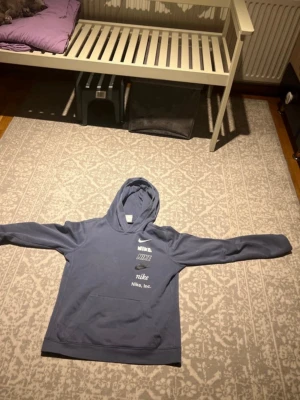 Blå hoodie från Nike med tryck - Snygg blå hoodie från Nike med flera vita logotyper och text på bröstet. Klassisk känguruficka framtill och huva. Tillverkad i mjukt material som känns skönt mot huden. Perfekt för en avslappnad och sportig stil.