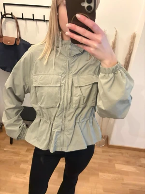 Jacka från Monki  - Vindjacka från Monki. Den har en huva med resår, resår vid ärmslut och reglerbar midja med dragsko för en anpassningsbar passform. Använd 1 gång. Färgen är dimgrön 