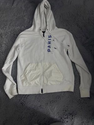 Vit PSG x Jordan hoodie med dragkedja - Säljer en vit hoodie från Jordan x PSG med dragkedja framtill och stor ficka. Trycket 'PARIS' i blått på bröstet och detaljer med både Jordan-loggan och PSG-emblemet. Tillverkad i mjukt bomullsmaterial och har justerbar huva med snören. Helt ny den är aldrig använd priset kan diskuteras