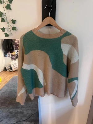 Beige och grön stickad tröja - Stickad tröja med abstrakt mönster i beige, grön och vit. Tröjan har rund halsringning och långa ärmar. Mjuk och skön passform, perfekt för lager på lager. Ingen synlig logga eller märke.