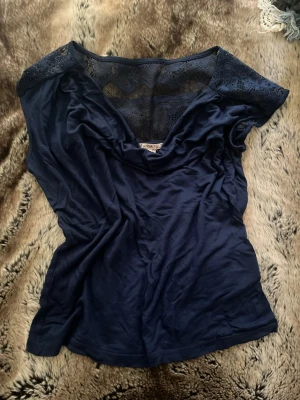 Mörkblå topp med spetsdetaljer - Mörkblå topp med v-ringning och snygga spetsdetaljer över axlar och rygg. Toppen har en feminin passform och är tillverkad i mjukt material som känns skönt mot huden. Perfekt för dig som gillar stilrena plagg med en twist.