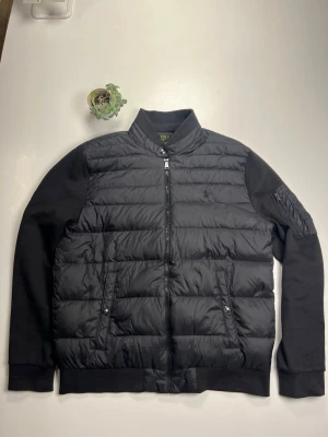 Svart cardigan Polo Ralph Lauren  - Svart cardigan  från Polo Ralph Lauren med quiltad framsida och rygg, samt släta ärmar i bomull. Jackan har dragkedja framtill och en cool ficka med dragkedja på ena ärmen. Insidan har ett rutigt foder. Perfekt för dig som gillar stilren och klassisk look. ❄️☔️