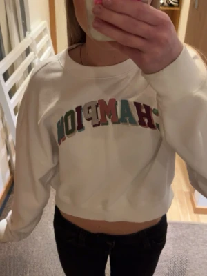 Vit croppad sweatshirt Champion - Säljer en vit croppad sweatshirt från Champion med broderad text i flera färger på bröstet. Tröjan har lång ärm och en avslappnad passform, perfekt till jeans eller tights. Superfräsch och trendig stil! Står att den är i storlek M men skulle definitivt säga att den är mer XS/S