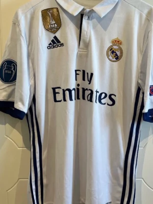 Real Madrid matchtröja Modrić 19 - Säljer en vit Real Madrid matchtröja från Adidas med Modrić och nummer 19 på ryggen. Tröjan har klubbmärke, sponsorlogga och Champions League-patchar på ärmarna. Klassisk pikékrage och blå detaljer. Perfekt för fotbollsfans!