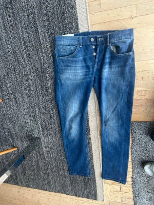 Blå jeans från dondup  - Säljer ett par blå jeans med knappgylf och klassisk femficksdesign. Jeansen har en snygg tvätt med slitningar och kontrastsömmar. Bakfickan har en diskret broderad detalj och det finns en röd märkning i linningen. Perfekta till vardagslooken. Dondup jeans George storlek 32 mycket bra skick lite kortare i längden 