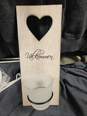 Välkommen skylt med ljushållare - Dekorativ skylt i trä med vitlaserad framsida och utskuret hjärta upptill. Texten 'Välkommen' är tryckt på framsidan och en frostad glasljushållare sitter fast med metallring. Baksidan är träfärgad med upphängningsanordning. Perfekt som inredningsdetalj.