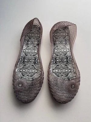 Genomskinliga ballerinaskor med blommor - Säljer ett par genomskinliga ballerinaskor i plast med små silverprickar och en söt blomdetalj framtill. Innersulan har ett zebramönster i svartvitt som ger en cool kontrast. Perfekta för dig som vill sticka ut med din stil!