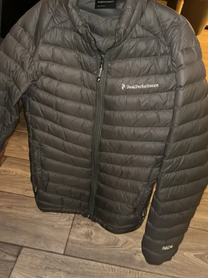 Svart dunjacka från Peak Performance - Snygg mörkgrön  dunjacka från Peak Performance med quiltad design och dragkedja framtill. Jackan har diskret logga på bröstet och har en modern, sportig look. Säljer denna för att jag behöver bli av med jackor då jag har för många. Nypris 1000kr