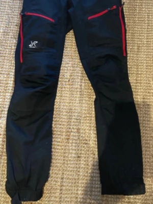 Svarta cargobyxor från RVRC M/38 - Säljer ett par svarta cargobyxor från RVRC, modell GPx Pro Pants. Byxorna har röda dragkedjor på fickorna och förstärkta partier vid knän och benslut. Tillverkade i slitstarkt material, perfekta för äventyr och outdoor.
