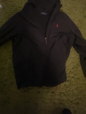Svart vindjacka från Polo Ralph Lauren - Snygg svart vindjacka från Polo Ralph Lauren med dragkedja framtill och den klassiska röda loggan broderad på bröstet. Jackan har en enkel och stilren design, perfekt för dig som gillar tidlös stil.