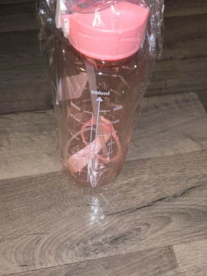 Rosa vattenflaska med måttskala - Transparent vattenflaska i plast med rosa lock och bärrem. Flaskan rymmer upp till 900 ml och har tydliga måttangivelser i både ml och oz på sidan. Praktisk för träning, skola eller utflykter.