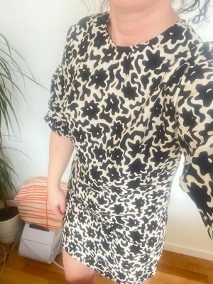 Kort blommig klänning i svart och beige - Säljer en kort klänning med svart och beige blommigt mönster. Klänningen har puffiga korta ärmar och rund halsringning. Perfekt för dig som gillar statement-plagg med coola detaljer och unik print.